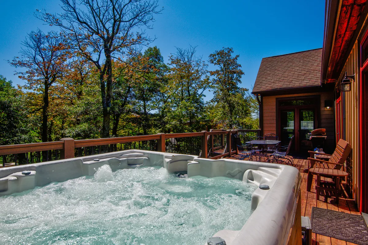 A tranquil view of a wooden deck nestled among tall trees, featuring a large hot tub, comfortable outdoor seating, and serene natural surroundings. The space is ideal for unwinding after outdoor adventures, enjoying morning coffee, or taking in sunset and starlit views from the tub.