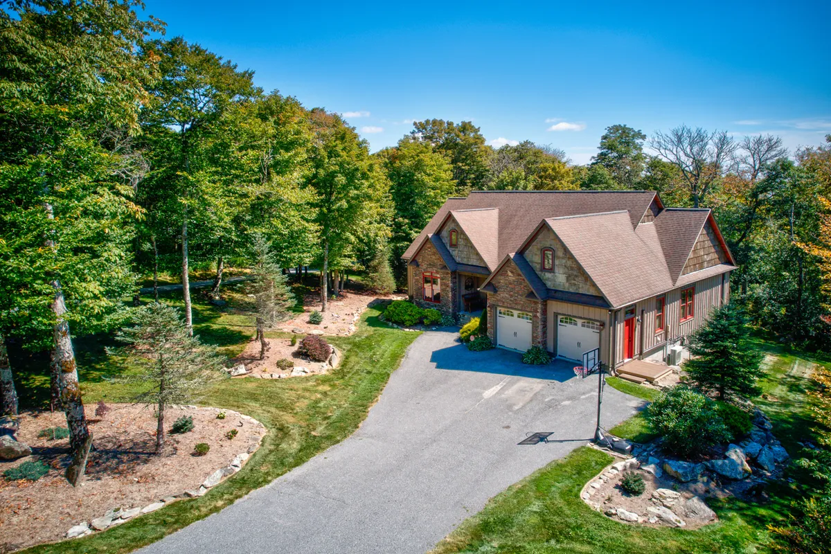 The image features the house nestled amid a beautifully landscaped garden and spacious driveway. The setting is peaceful and inviting, with hints of nearby nature trails and ample space for hosting outdoor activities and gatherings.
