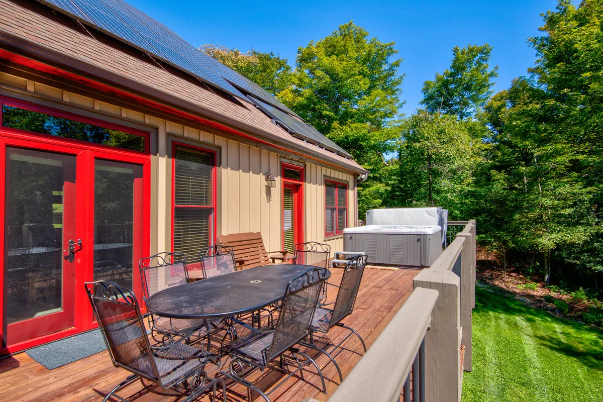 This image features a spacious deck with a dining table, a large hot tub, and vibrant red doors, all surrounded by lush green trees. The scene captures a group of people enjoying outdoor dining, relaxing in the hot tub, and gathering with friends, set against a beautiful sunset backdrop. Solar panels on the roof add an eco-friendly aspect, and the atmosphere is lively and welcoming.