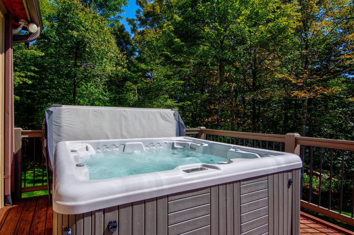 This exterior image features a spacious deck with a hot tub nestled among tall, leafy trees. The serene setting is perfect for relaxation, with the hot tub providing a direct connection to the outdoors and the surrounding natural beauty. The view suggests a peaceful mountain retreat atmosphere, inviting guests to unwind while immersed in nature.