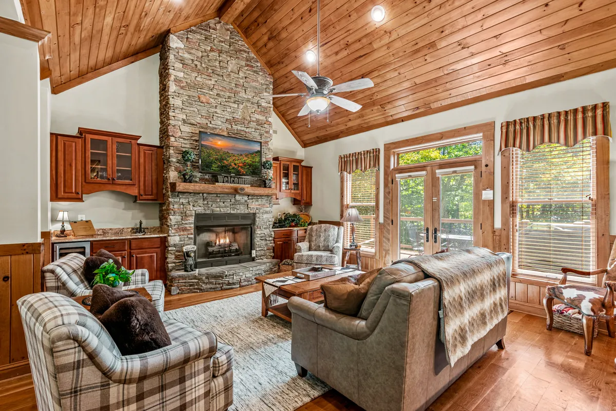 This image features a warm, inviting living room with a prominent stone fireplace, a wooden ceiling, comfortable seating arranged for group gatherings, and large windows letting in abundant natural light. The view through the windows alludes to the surrounding landscape, while the layout highlights the open-plan nature of the space.
