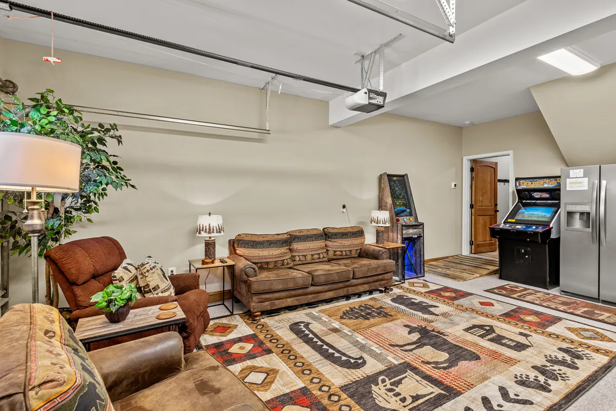 A cozy corner of the game room featuring a sofa, an arcade machine, a small refrigerator, and a potted plant. The relaxed setup invites guests to lounge, grab drinks, and enjoy arcade classics in a comfortable, informal setting.