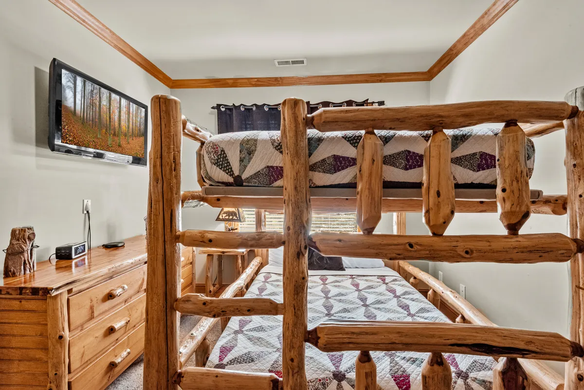 A family-friendly bedroom featuring a sturdy log-frame bunk bed, cozy quilts, a wall-mounted TV, and a wooden dresser. The atmosphere is both playful and relaxing, ideal for children or group stays.