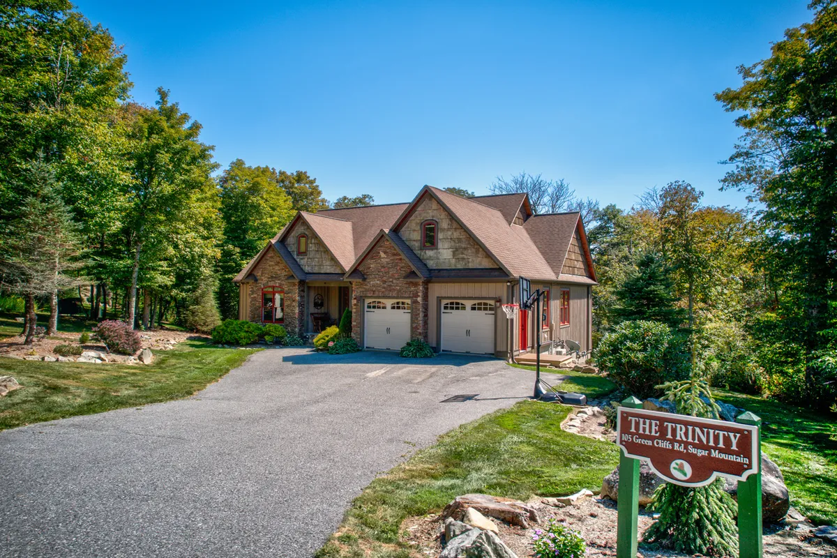 The welcoming stone facade, well-maintained driveway, lush landscaping, and prominent 'THE TRINITY' sign create a sense of arrival. The covered front porch suggests enjoying morning coffee, and the inviting entrance sets an upscale, comfortable tone.