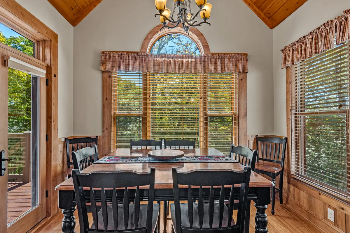 A formal dining area with a large wooden table under a chandelier, surrounded by large windows and wooden accents. The setting is inviting and bright, with natural light and views that connect indoor dining with the surrounding landscape.