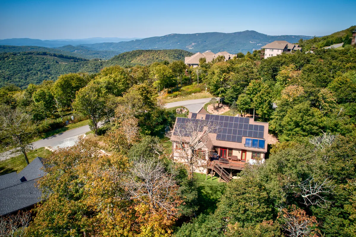 This image features the exterior of the house with visible solar panels integrated into the roof, surrounded by trees and backed by majestic mountains. The lighting suggests a serene sunset, and the porch invites relaxation while connecting guests to the stunning natural landscape.