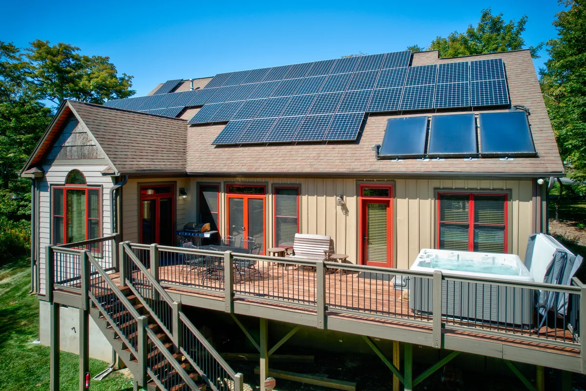The exterior deck showcases a hot tub, deck seating, and solar panels on the roof. The space feels inviting for both daytime and evening leisure, with trees providing privacy. The red window frames add a designer touch, and the hot tub is central for relaxation and stargazing.