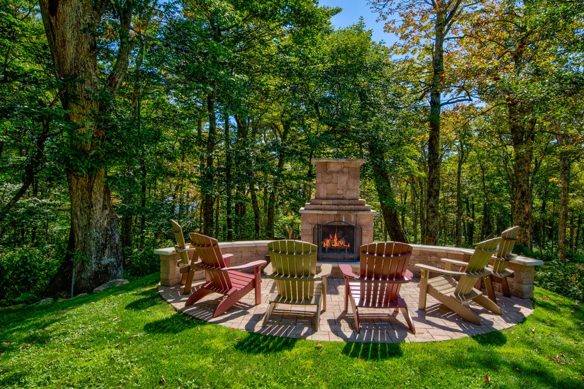 A cozy stone outdoor fireplace with Adirondack chairs arranged in a circle, set on a rustic stone patio surrounded by nature. The space invites guests to gather, make s'mores, and enjoy stories or stargazing under the night sky. The warm glow of the fire enhances the inviting atmosphere.