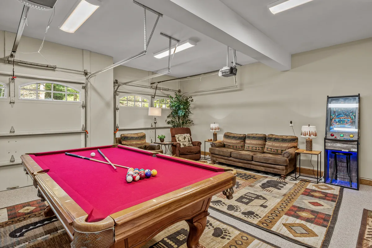 A vibrant game room featuring a pool table as the central attraction. To one side is a comfy seating area with an indoor plant for a homely touch, and an arcade machine stands nearby, adding a nostalgic, retro vibe. The open layout suggests ample space for multiple activities and social gatherings.